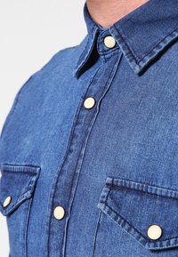 Džínová košile v tmavě modré barvě s špičatým límcem, předním zapínáním na knoflíky a dvěma náprsními kapsami se žlutými knoflíky a jemnou texturou.