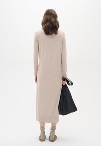 Beige pletené šaty s dlouhými rukávy, úzkým střihem a bočním rozparkem; spárováno s béžovými ponožkami a držící černou koženou tašku.