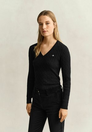 Jeune femme aux longs cheveux blonds porte un pull noir à col en V en maille torsadée et un pantalon noir, debout devant un fond clair uni.