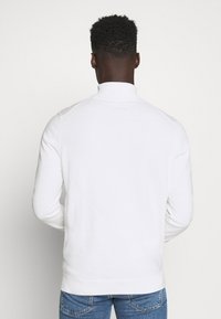 Uomo di spalle che indossa un maglione bianco a maniche lunghe e jeans blu su uno sfondo bianco semplice.