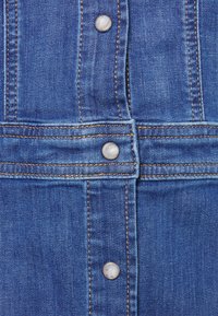 Denim w średnim niebieskim kolorze z widocznym szwem, wyposażony w metalowe zatrzaski w kolorze srebrnym oraz gładką fakturę. Prosty krój.