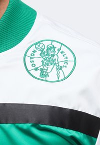 Λογότυπο των Boston Celtics με έναν λεπτποχάλκινο να γυρίζει μια μπάλα μπάσκετ, κεντημένο σε λευκό, πράσινο και μαύρο αθλητικό τζάκετ.