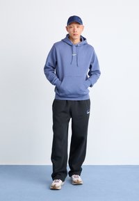 Felpa blu con tasca frontale e logo verde, abbinata a pantaloni jogger neri con logo blu e scarpe da ginnastica beige con accenti rossi.