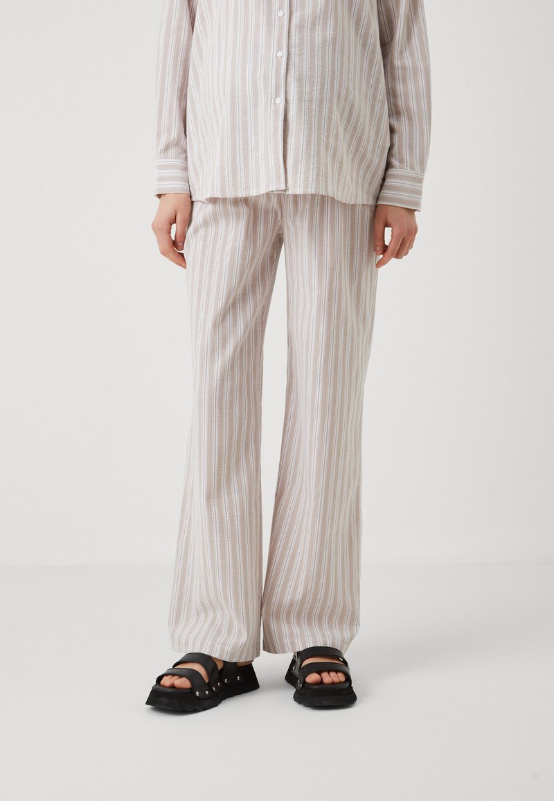 Ensemble de loisirs rayé beige comprenant une chemise boutonnée et un pantalon évasé. Le tissu a une texture douce avec de fines lignes blanches. Porté avec des sandales noires.
