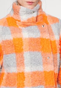 Farverig ternet frakke lavet af blød, fuzzy stof med orange, grå og hvide firkanter. Høj krave med metaltrykknapsdetaljer.
