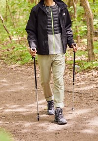Personne marchant sur un sentier forestier avec des bâtons de trekking, vêtue d'une veste noire, d'un pantalon beige et de chaussures de randonnée.