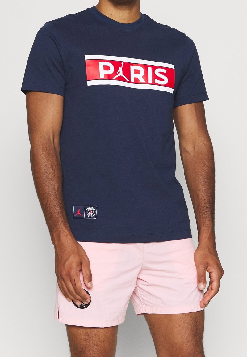 Námořnická modrá tričko s červeným a bílým grafickým prvkem "PARIS" a logem Jumpman, vyrobené z bavlny. Nosené s světle růžovými šortkami.