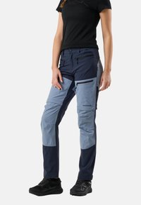 Vrouw draagt marineblauwe en lichtblauwe outdoorbroek met zwarte sneakers, staand tegen een witte achtergrond.