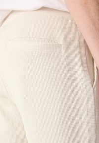 Pantaloni beige con texture a righe, dotati di una tasca posteriore e una cintura liscia. Vista ravvicinata che mette in evidenza il materiale e i dettagli del design.