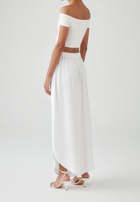 Witte off-shoulder crop top gecombineerd met een witte maxi-rok met een elastische tailleband en hoge split. Strappy witte hakken.