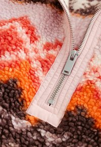 Polarowy pulower z żywymi pomarańczowymi, różowymi i brązowymi wzorami. Posiada srebrny zamek i teksturowany, puszysty materiał.