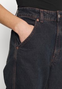Main reposant dans la poche avant d'un jean noir délavé foncé avec des surpiqûres orange visibles et un haut noir rentré à l'intérieur.