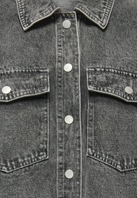 Gros plan sur le devant d'une veste en denim noir montrant le col, les boutons-pression en métal, et deux poches poitrine à rabat avec fermeture à bouton.