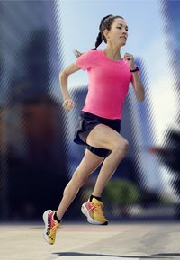 Coureur en t-shirt ajusté rose vif et short noir. Porte des sneakers jaunes avec des accents rouges. Arrière-plan avec un cadre urbain flou.