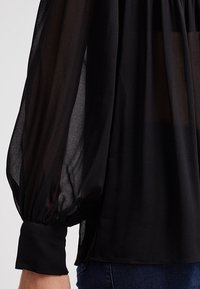 Blusa nera trasparente con maniche lunghe a sbuffo. Presenta un polsino aderente e una texture leggera e semi-trasparente.