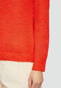 Strickpullover in leuchtendem Rot mit strukturierter Oberfläche, ausgestattet mit gerippten Bündchen und einer V-förmigen Nahtdetail. Getragen mit hellen Hosen.