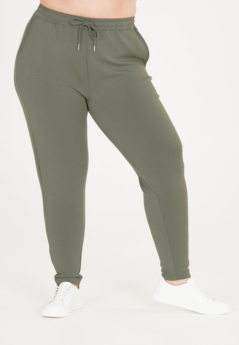 Olivengrønne joggerbukser med sidelommer og elastisk talje med snor. Stoffet fremstår blødt med en glat struktur. Sammen med hvide sneakers.