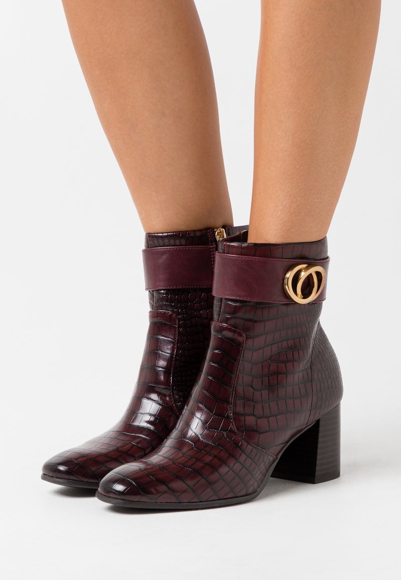 Bottines bordeaux à texture crocodile avec talons blocs et détail de boucle double anneau doré portées sur jambes nues.