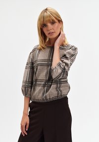 Blondynka ubrana w szary top w kratkę z bufkami i czarne spodnie, delikatnie dotykająca włosów, na tle jednolitego białego tła.