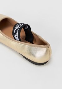 Guldballerinaskor med en glänsande metallisk finish, med en svart elastisk rem märkt "MOSCHINO" och en svart gummisula.