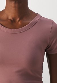 Korte mouwen, aansluitend top in zachte mauve stof. Heeft een ronde halslijn en een gladde textuur met minimale naden.