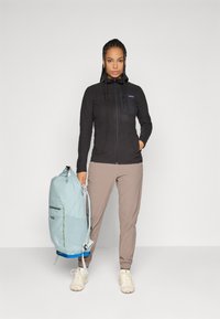 Woman with braided hair wearing black zip-up jacket, beige pants, and white sneakers, holding a large light blue backpack by her side.