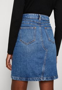 Kvinna som bär en blå denimkjol med hög midja, bakfickor och bältesloopar, i kombination med en långärmad svart topp.