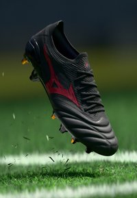 Schwarzer Fußballschuh mit rotem Logo, in der Luft über einem Rasenfeld mit weißer Linie, der kleine Grasstücke aufwirbelt.