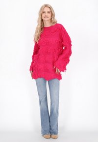 Roze gebreide trui met franjes en lange mouwen, gecombineerd met lichtblauwe flare jeans en beige schoenen.