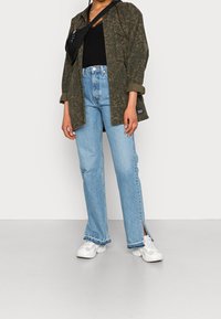 Chemise à motif vert olive, haut noir, jeans droit bleu clair avec fente sur les côtés, et baskets blanches. Design simple avec coupe décontractée.