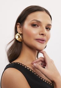 Złote, rzeźbiarskie kolczyki o gładkiej fakturze, w kształcie organicznym. Noszone przez osobę w czarnej bluzce z falbaniastym dekoltem.