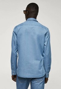 Camicia in denim azzurro chiaro con maniche lunghe, polsini bottonati, colletto classico e una texture liscia; la vista posteriore mostra cuciture dritte e un orlo curvo.
