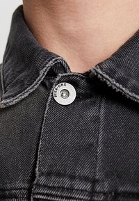 Cuello de chaqueta de mezclilla negra con un botón plateado con la inscripción "PIER.ONE". Costuras visibles y detalles de tela texturizada.