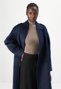 Cabană din lână bleumarin cu guler mare și nasturi, asortată cu un pulover turtleneck moltonat de culoare neutră și pantaloni negri, creând un look stratificat.