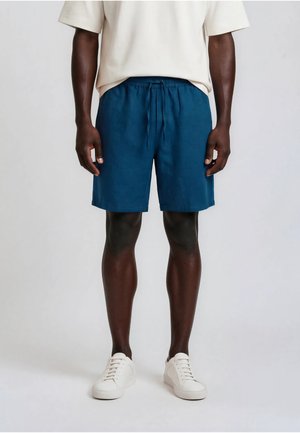 Homme portant un short bleu à cordon, une chemise blanche à manches courtes et des baskets blanches à lacets, debout devant un fond clair uni.