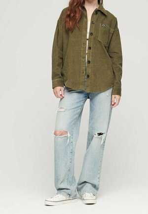 Mujer con una camisa de pana de color verde oliva de gran tamaño abotonada, vaqueros azul claro desgarrados y zapatillas blancas, de pie contra un fondo liso.