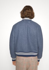 Blå ullbombarjacka med ribbad vit och marinblå randig krage och manschetter. Houndstooth-mönstrade byxor delvis synliga.