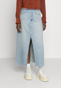 Jupe maxi en denim bleu clair avec une fente frontale, un ourlet effiloché et des passants de ceinture ; assortie à des baskets blanches et des chaussettes tube.