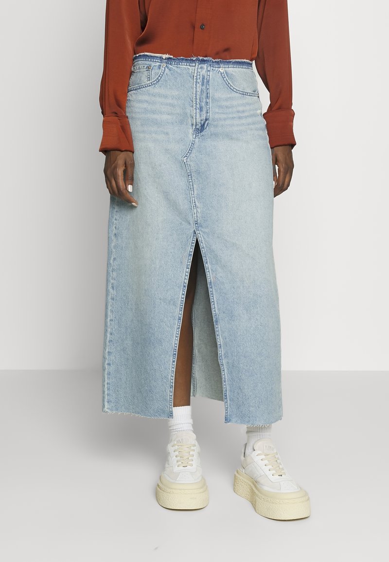Jupe maxi en denim bleu clair avec une fente frontale, un ourlet effiloché et des passants de ceinture ; assortie à des baskets blanches et des chaussettes tube.