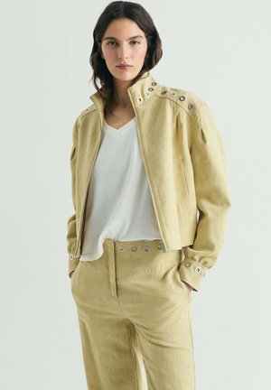Mujer con cabello castaño lleva chaqueta y pantalones beige con detalles de ojales metálicos, combinados con una camiseta blanca de cuello en V, de pie con las manos en los bolsillos.