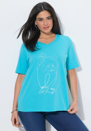 Donna con capelli lunghi e scuri che indossa una maglietta blu brillante con un disegno bianco a linee di un orso e la scritta "migliori amici", abbinata a jeans scuri.