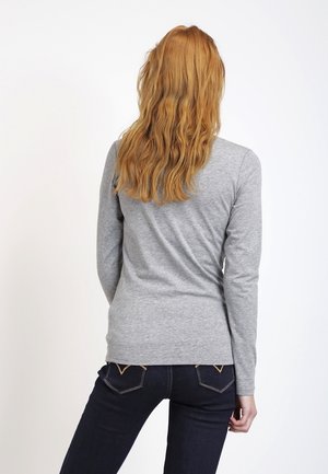 Femme aux longs cheveux roux ondulés, portant un t-shirt gris à manches longues et un jean bleu foncé, debout de dos sur fond blanc.