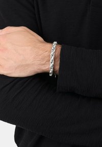 Pulsera de cadena de plata en una muñeca, con eslabones entrelazados y un acabado pulido. Lleva puesta sobre una manga negra texturizada.