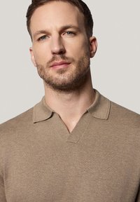 Beżowa dzianinowa koszula polo z dekoltem w serek i krótkim kołnierzykiem. Materiał ma gładką teksturę i dopasowany krój.