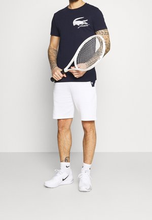 Marineblaues T-Shirt mit weißem Lacoste-Logo, weiße Shorts, weiße Nike-Schuhe, hält einen weißen Tennisschläger und steht vor einem hellgrauen Hintergrund.