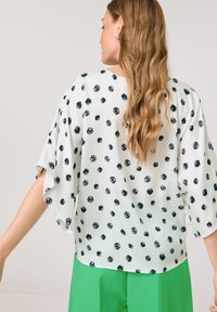 Weiße Bluse mit schwarz-grünen, kreisförmigen Mustern, kurzen Ärmeln und lockerer Passform, kombiniert mit leuchtend grünen Hosen. Glatte Textur.