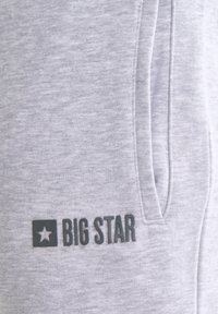 Tessuto grigio chiaro con tasca verticale con cerniera e logo "BIG STAR" nero accanto a un simbolo a stella sul lato inferiore sinistro.