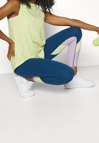 Světle zelený top bez rukávů, modré legíny s purpurovými a zelenými geometrickými vzory a bílé sportovní boty.