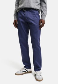 Lila Baumwollhosen mit geradem Bein, seitlichen Taschen und Gürtelschlaufen, kombiniert mit weißen und grauen Sneakers mit grünen Streifen.