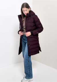 Giacca a vento viola scuro con cappuccio, chiusura lampo frontale e tasche laterali, indossata su un top marrone e jeans blu, esposta su uno sfondo neutro.